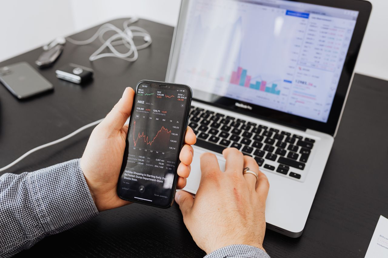 Personne analysant des données boursières avec un smartphone et un ordinateur portable, en intérieur.